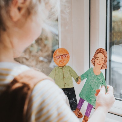 Ein kleines Mädchen steht am Fenster und hält zwei bemalte Pappfiguren in der Hand, eine Frau und einen Mann mit Bart und Brille.