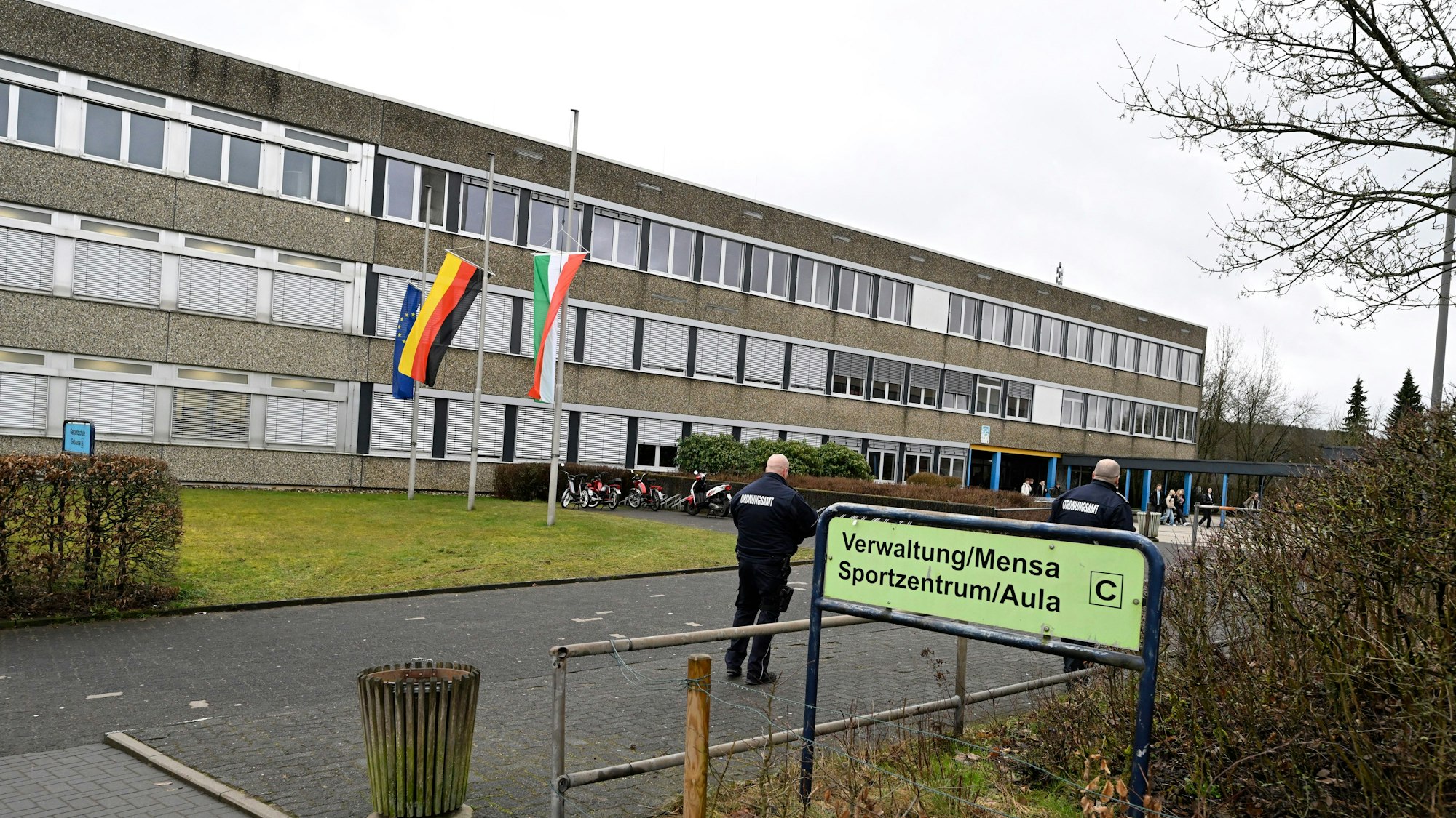 13.03.2023, Nordrhein-Westfalen, Freudenberg: Mitarbeiter des Ordnungsamtes stehen vor dem Schulzentrum des ermordeten Mädchens. Die Schule hat geöffnet, die Fahnen sind auf Halbmast geflaggt.
