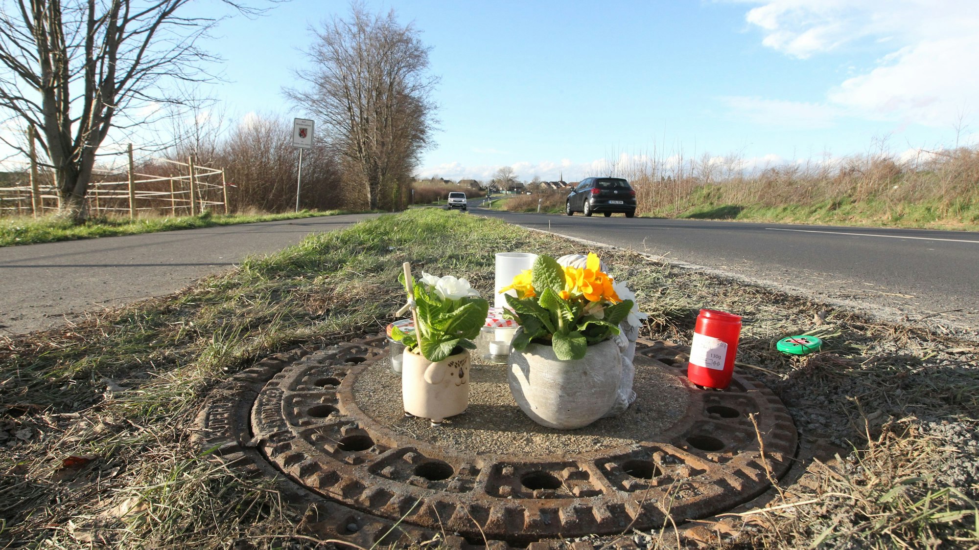Ein Gullydeckel am Rand einer Landstraße. Menschen haben Blumen und Kerzen daraufgestellt.