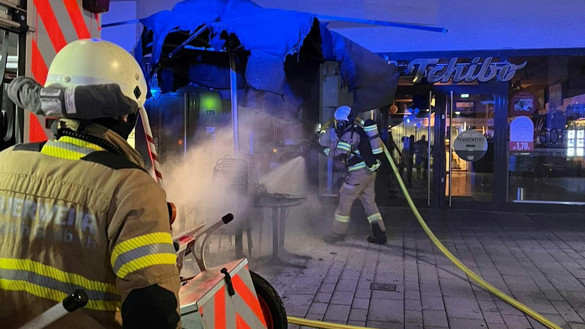 Ein Feuerwehrmann löscht einen rauchenden Schirm vor einem Geschäft, ein weiterer steht an einem Feuerwehrfahrzeug.