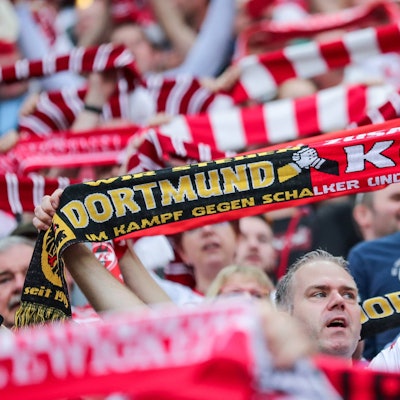 Ein Fußball-Fan hält einen Fan-Schal hoch, auf dem die Logos und Schriftzüge von Borussia Dortmund und dem 1. FC Köln zu sehen sind.