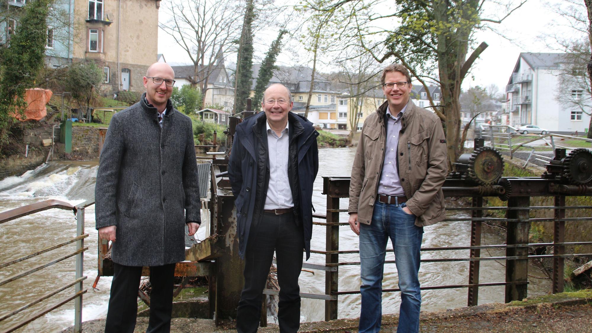 Die drei Männer sind vor einem Stauwehr zu sehen, dahinter das rauschende Wasser der Urft.