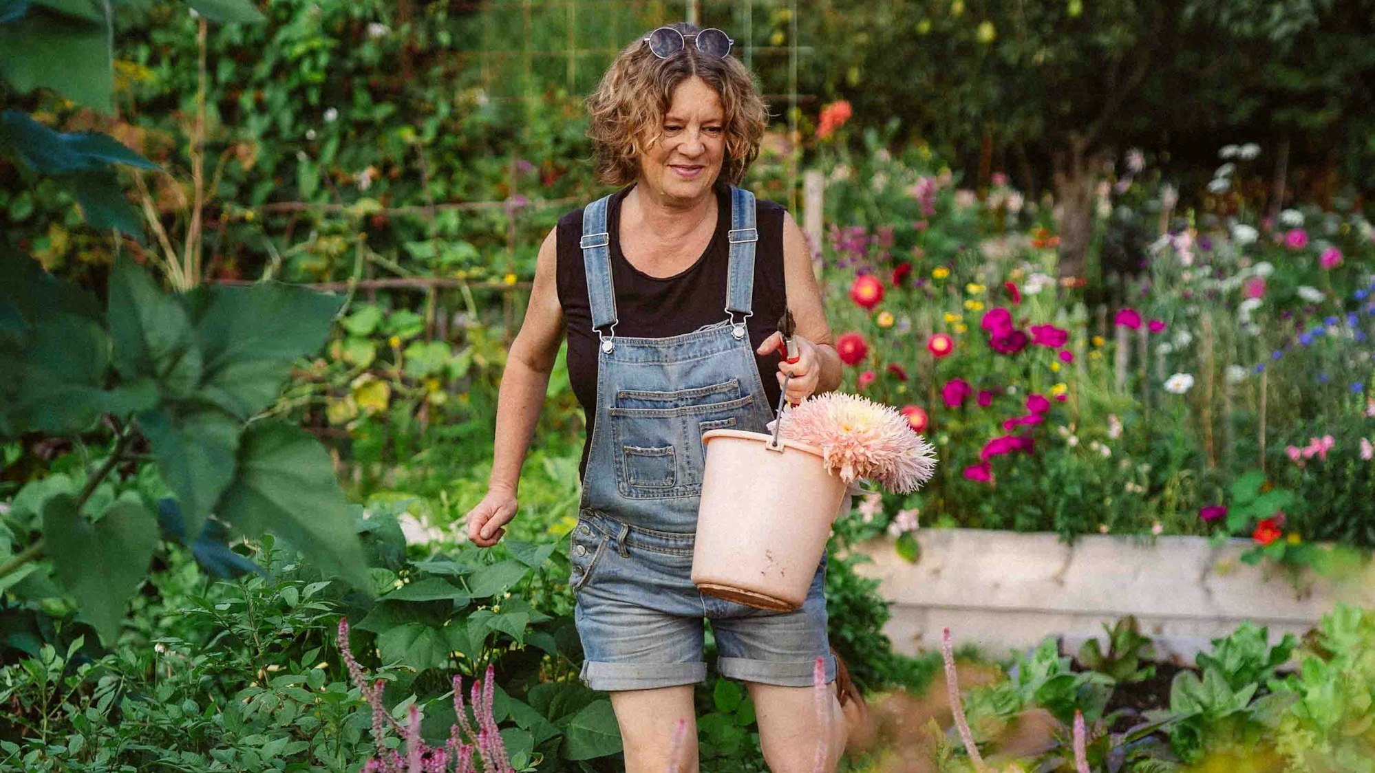 Anja Klein trägt eine Jeans-Latzhose und ein schwarzes T-Shirt und hält einen rosafarbenen Eimer mit Blumen in der Hand.