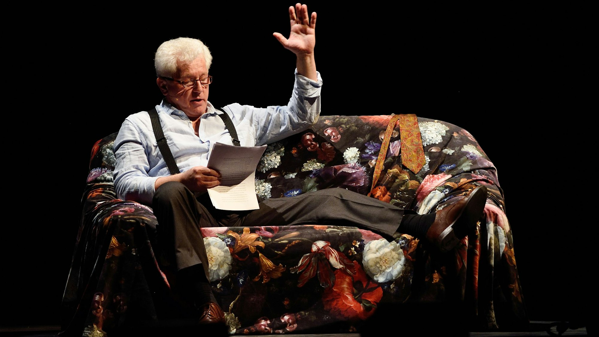 Schauspieler Miroslav Nemec sitzt auf einem Sessel auf der Bühne. Er hat ein Bein hochgelegt und liest von Zetteln. Mit der linken Hand gestikuliert er.