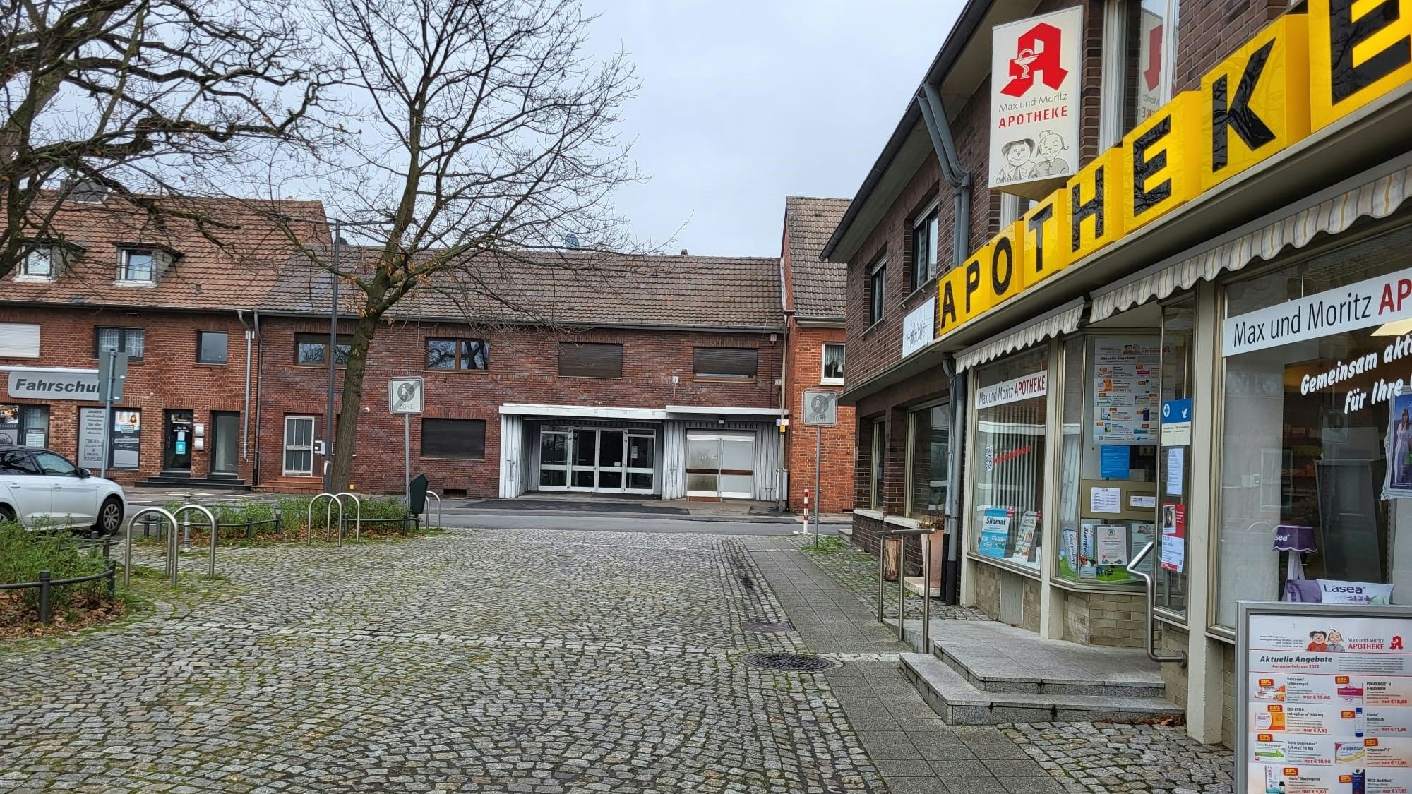 An einem gepflasterten Platz ist rechts eine Apotheke, im Hintergrund stehen Häuser aus Backstein.