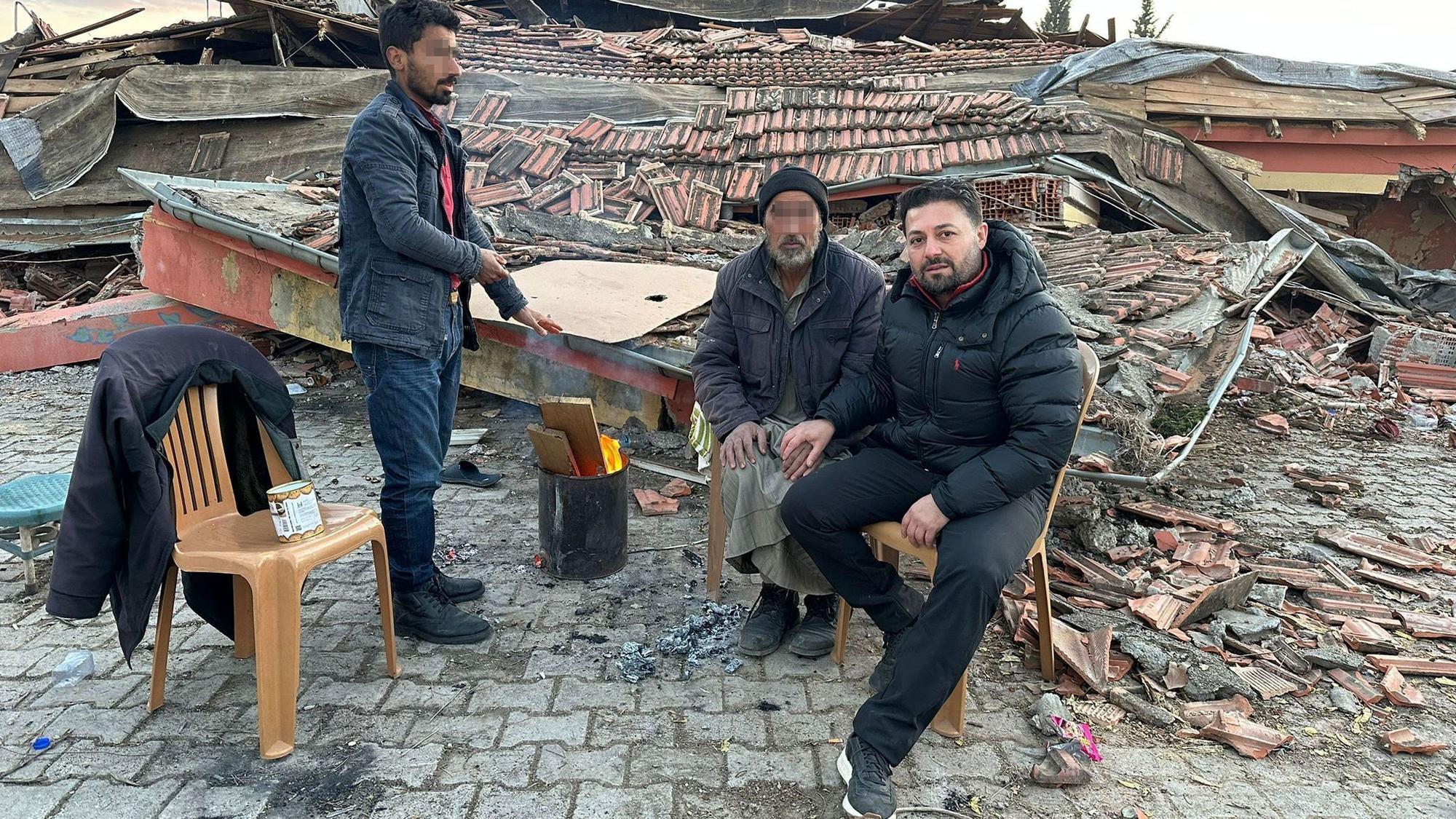 Der Euskirchener Ali Demirbas und zwei weitere Männer sitzen auf Plastikstühlen, um sie herum die Trümmer zerstörter Häuser.