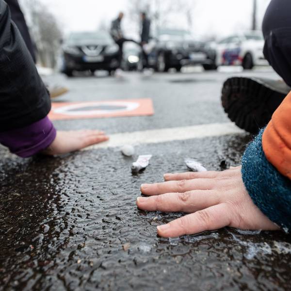 Klimaschutzaktivisten der „Letzten Generation“ kleben sich mit Sekunden kleben auf dem Asphalt einer Straße fest. Im Hintergrund stehen Autofahrer deswegen im Stau.