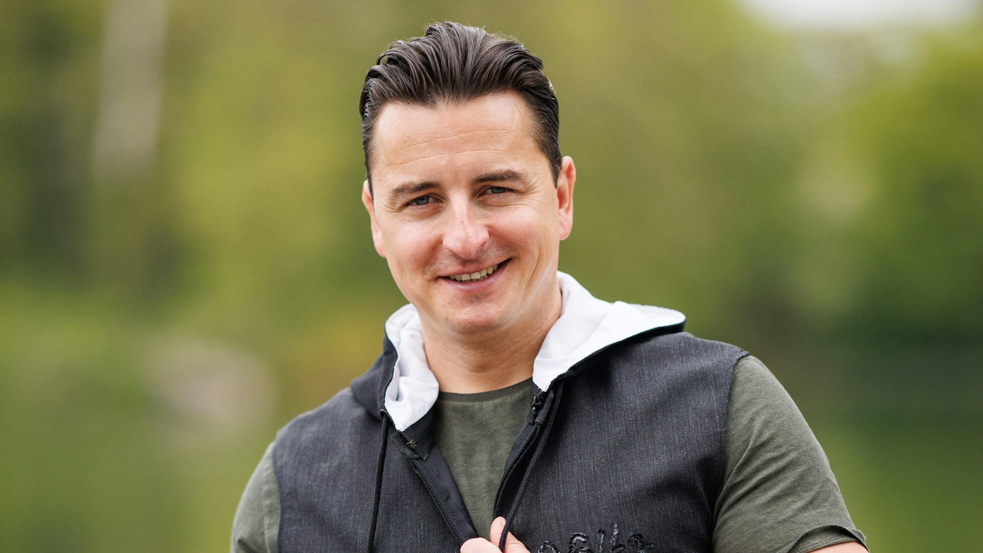 Andreas Gabalier lächelt vor grünen Bäumen in die Kamera.