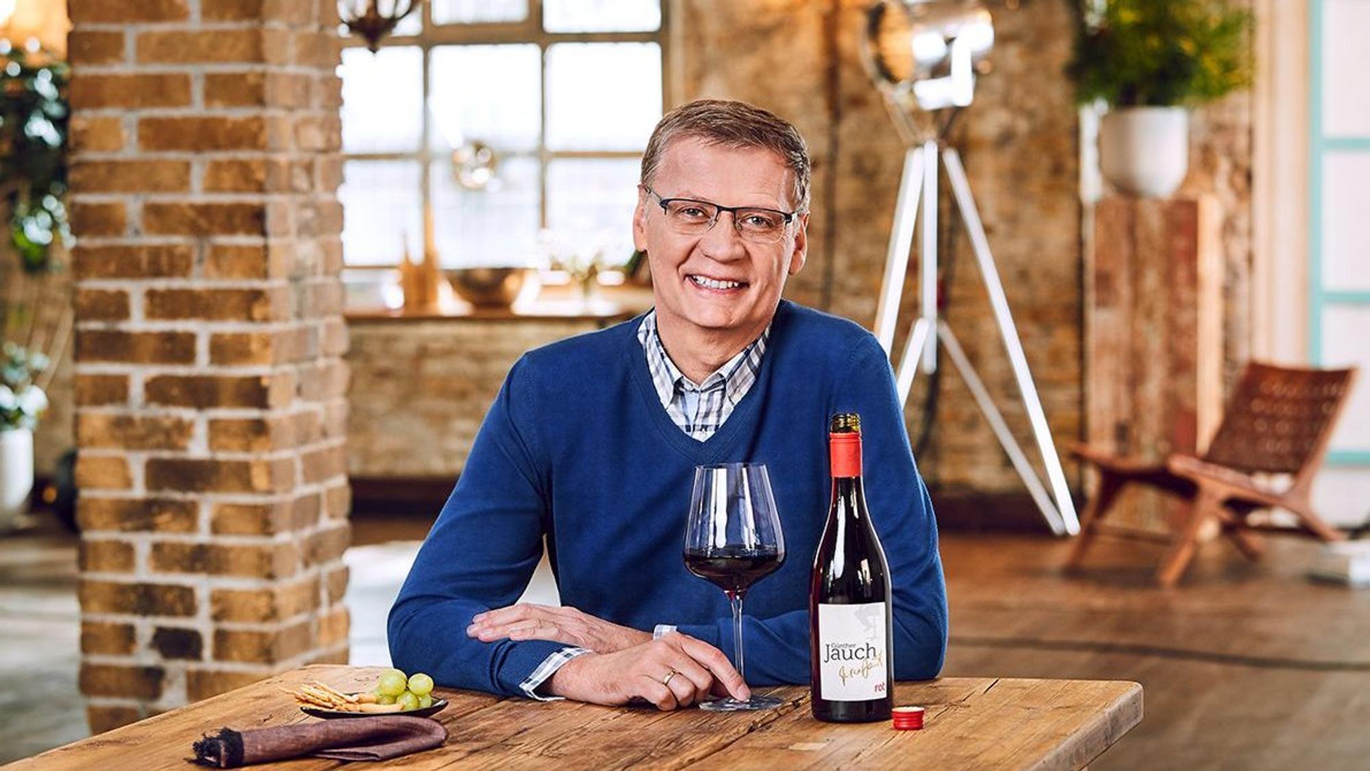 Günther Jauch sitzt mit einem Glas Wein an einem Tisch. Die offene Flasche steht daneben.