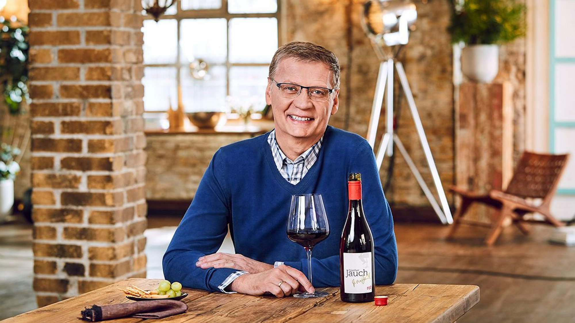 Günther Jauch sitzt mit einem Glas Wein an einem Tisch. Die offene Flasche steht daneben.