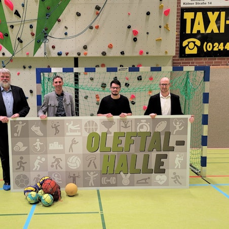 Die vier Männer stehen in einer Turnhalle und halten ein Plakat mit dem Namen in den Händen.
