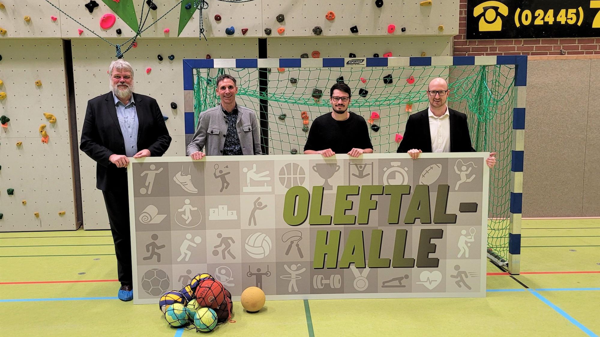 Die vier Männer stehen in einer Turnhalle und halten ein Plakat mit dem Namen in den Händen.