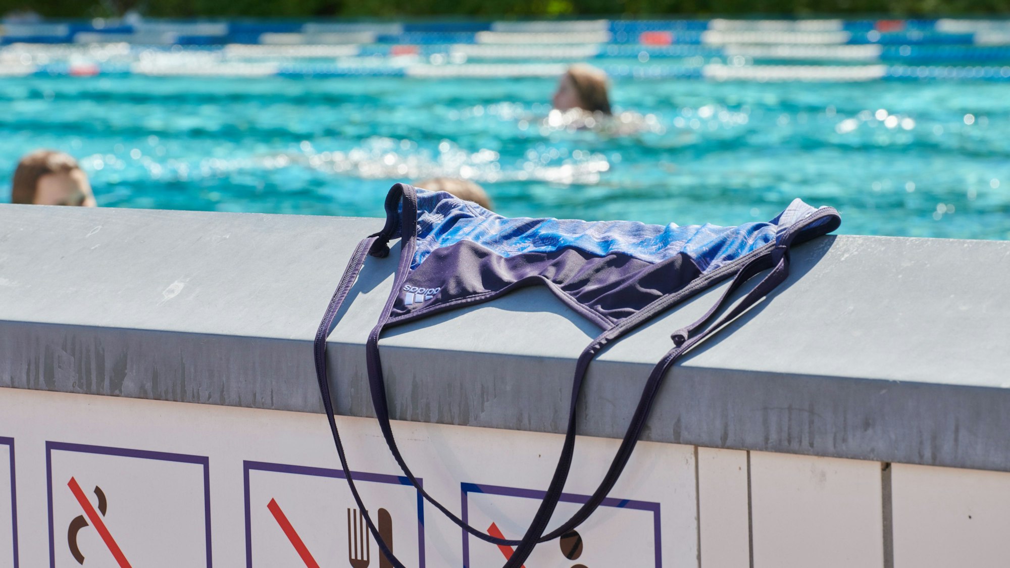 Ein Badeanzug liegt auf einer Mauer im Schwimmbad, dahinter schwimmen Menschen im Becken ihre Bahnen.