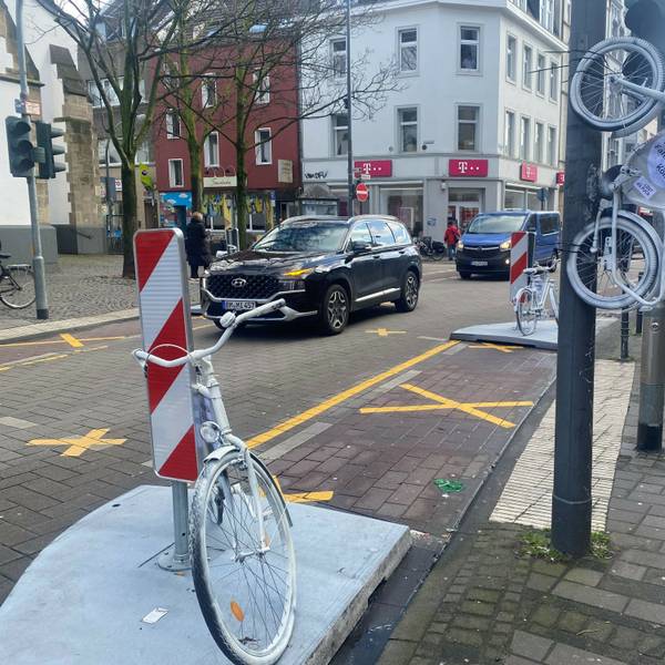Auf der Venloer Straße stehen zwei weiße Fahrräder am Straßenrand, ein drittes hängt an einem Laternenmast.
