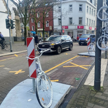 Auf der Venloer Straße stehen zwei weiße Fahrräder am Straßenrand, ein drittes hängt an einem Laternenmast.