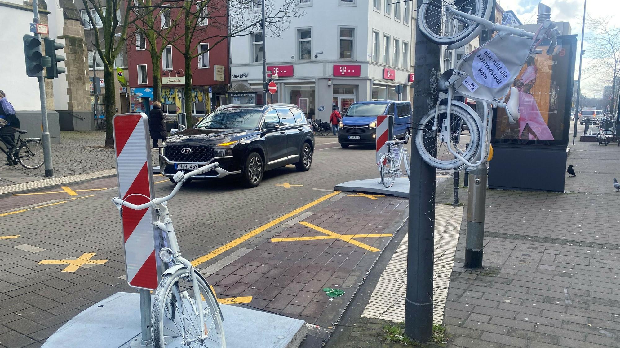 Auf der Venloer Straße stehen zwei weiße Fahrräder am Straßenrand, ein drittes hängt an einem Laternenmast.