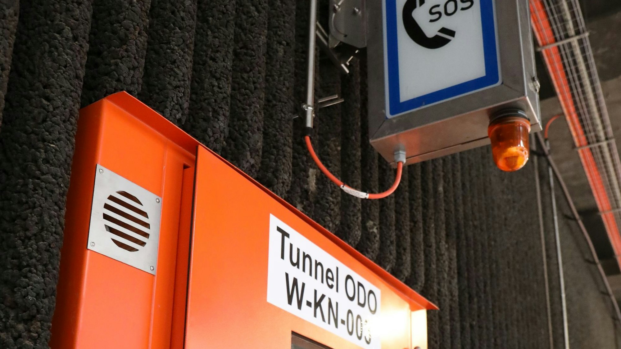 Über einer roten, in der Wand eingelassenen Stahltür hängt ein Schild mit dem Symbol eines Telefons und den Buchstaben SOS.