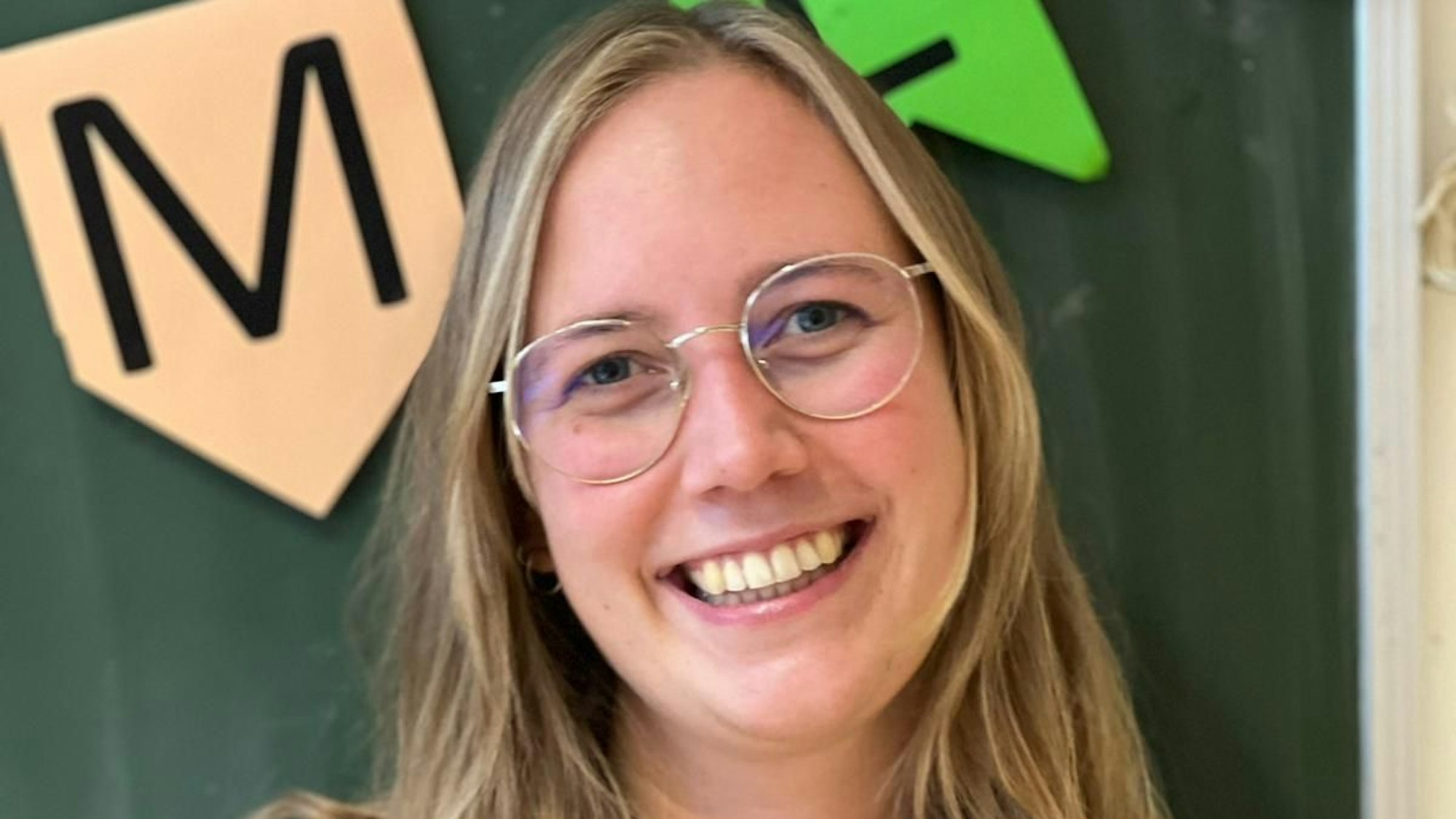 Eine blonde Frau mit Brille lacht in die Kamera. Hinter ihr sieht man eine Tafel mit Buchstaben.