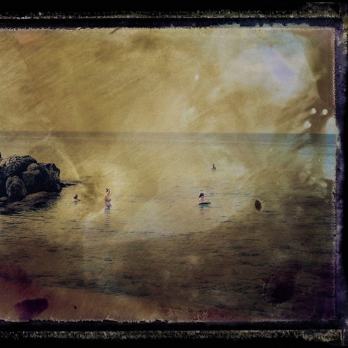 Die von braunen Schlieren durchzogene Fotografie zeigt eine Meeresbucht mit wenigen Badenden.