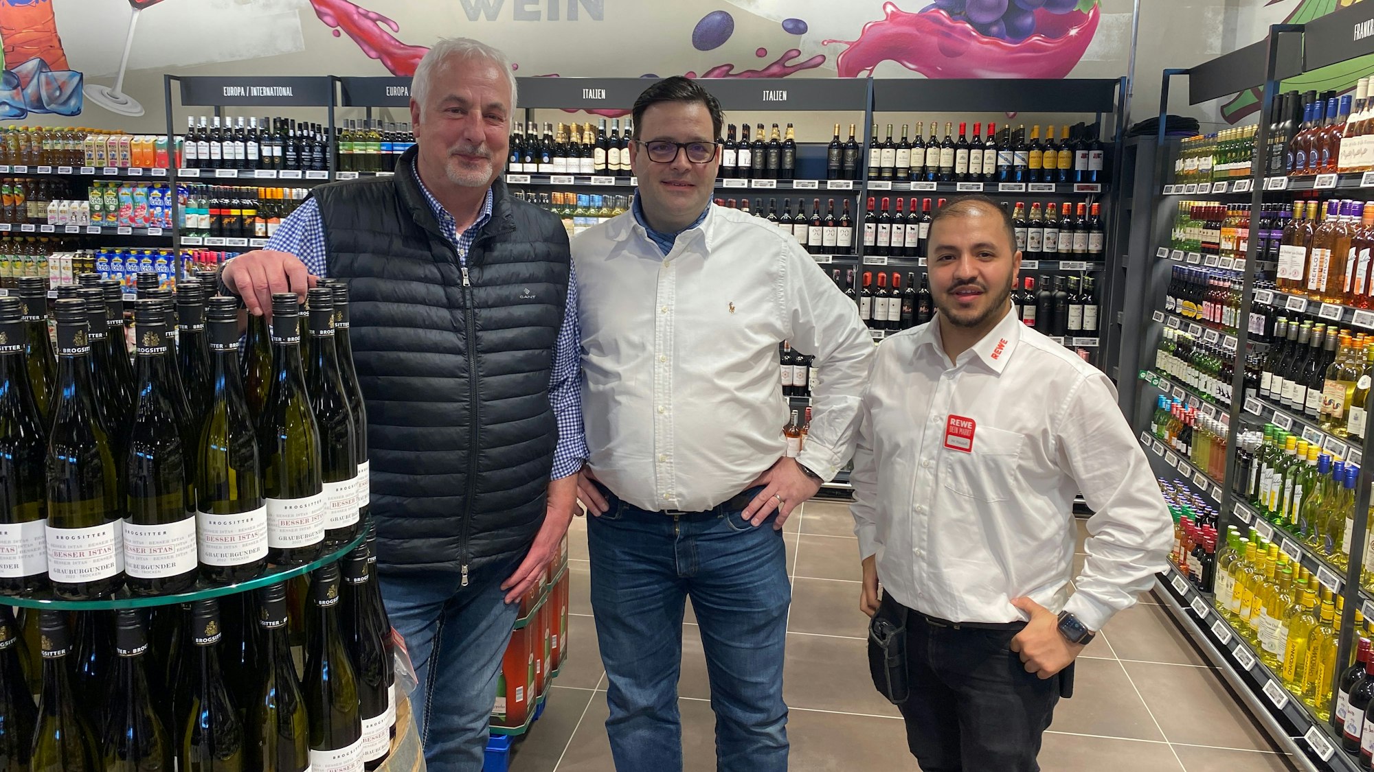 Die drei Männer stehen in der Getränkeabteilung neben einem Weinregal.
