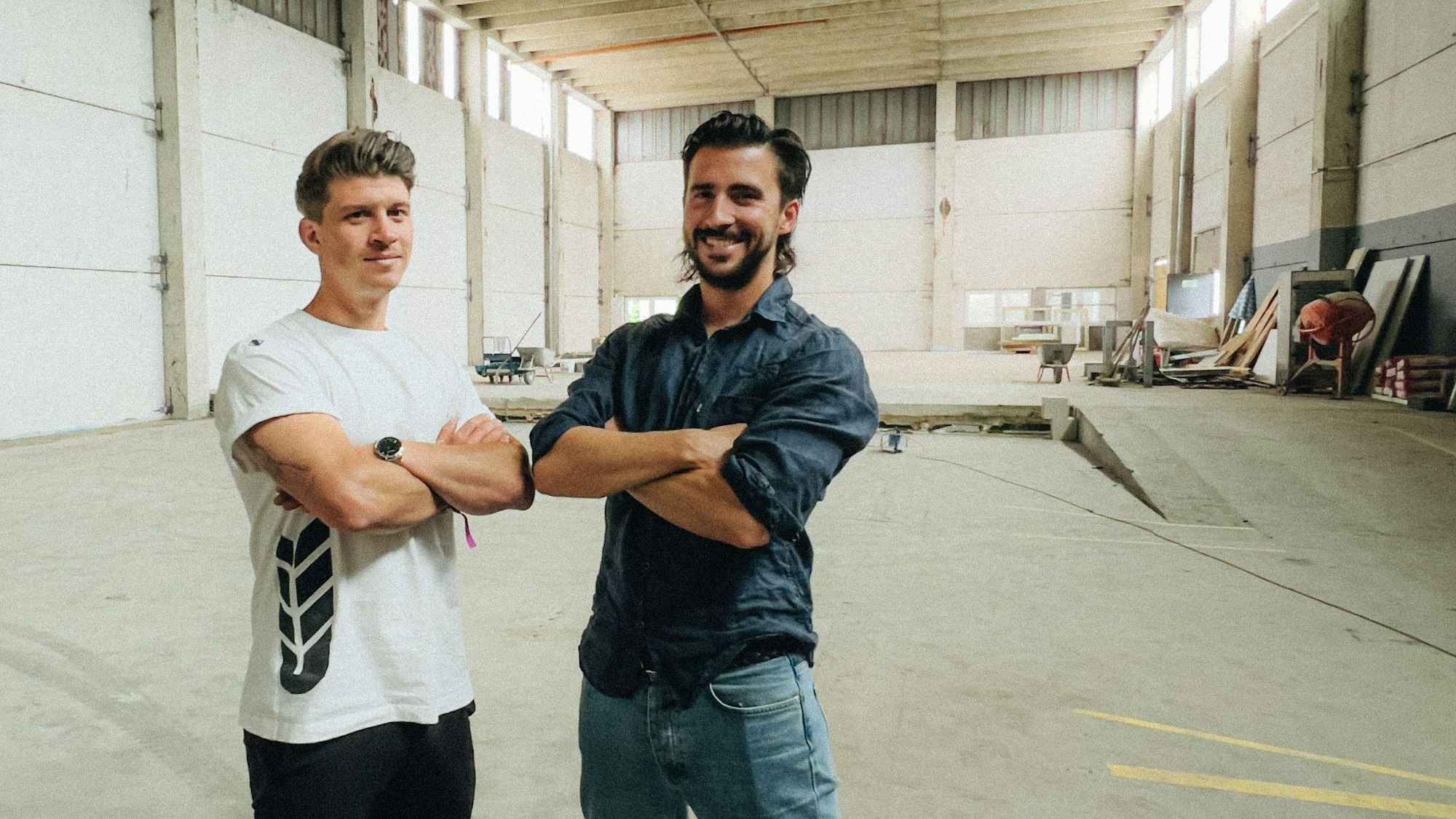Die Zappes-Gründer Nicolas Lutz und Maximilian Koeser posieren mit verschränkten Armen in einer Lagerhalle in Köln-Vogelsang.