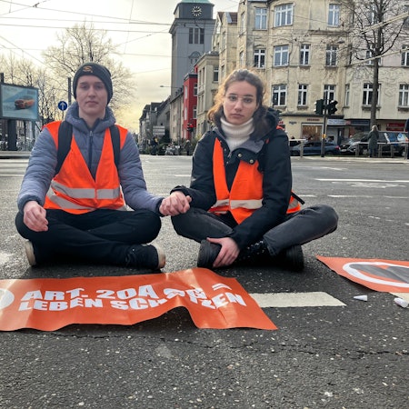 Ein Aktivist und eine Aktivistin sitzen Hand in Hand auf der Straße in Köln.