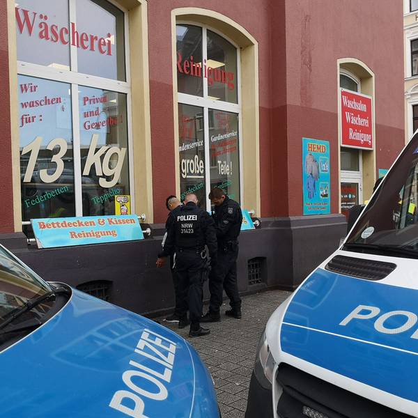 Zwei Polizisten durchsuchen einen Mann. Im Vordergrund sind zwei Polizei-Fahrzeuge zu sehen.