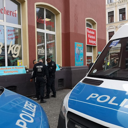 Zwei Polizisten durchsuchen einen Mann. Im Vordergrund sind zwei Polizei-Fahrzeuge zu sehen.