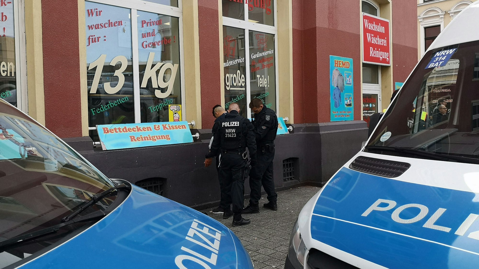 Zwei Polizisten durchsuchen einen Mann. Im Vordergrund sind zwei Polizei-Fahrzeuge zu sehen.