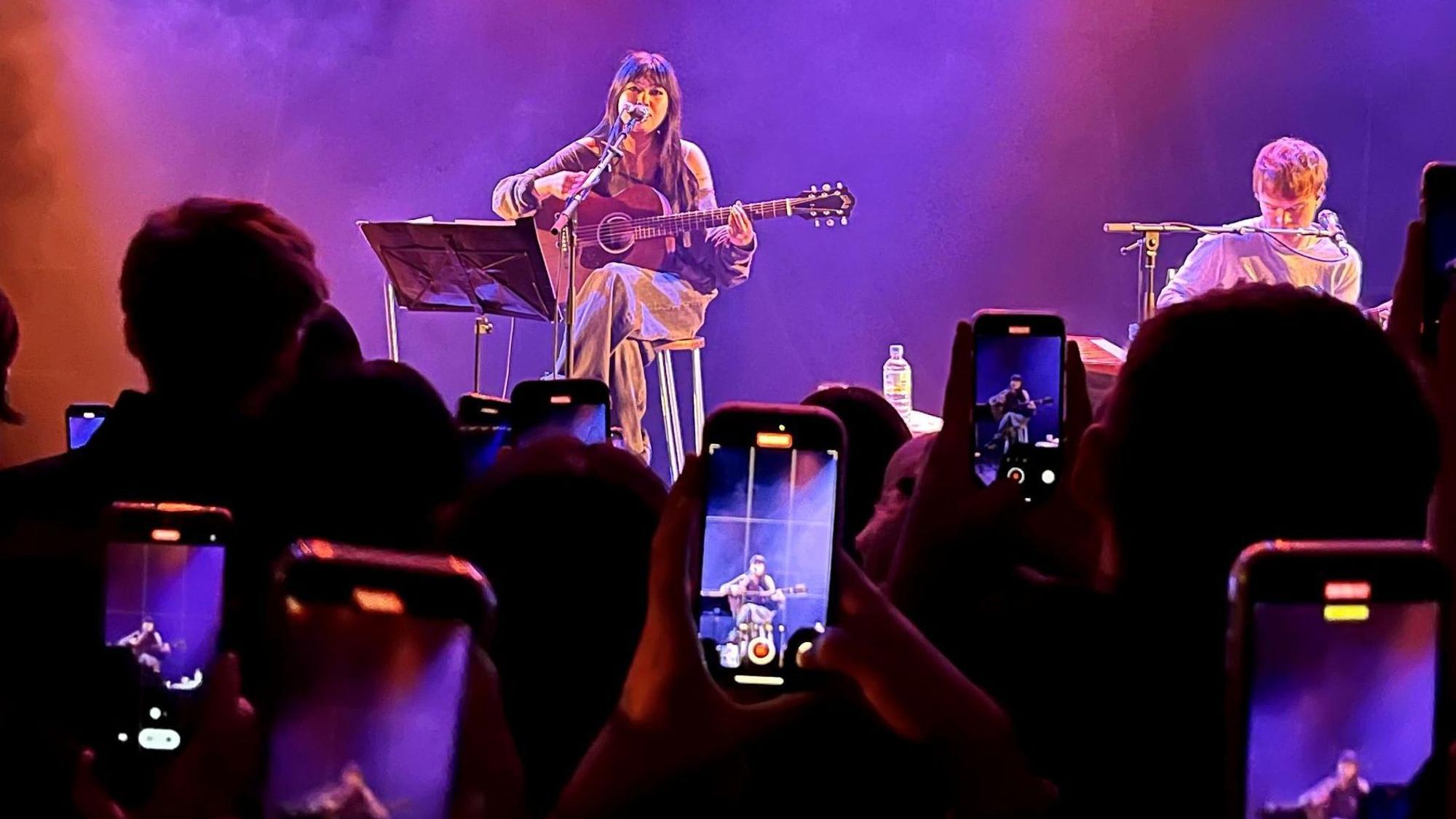 Die englische Sängerin Beabadoobee sitzt am 17. März 2023 mit einer Gitarre auf einem Barstuhl auf der Bühne des Kölner Bürgerhaus Stollwercks. Ihre Fans filmen sie mit ihren Handys ab.