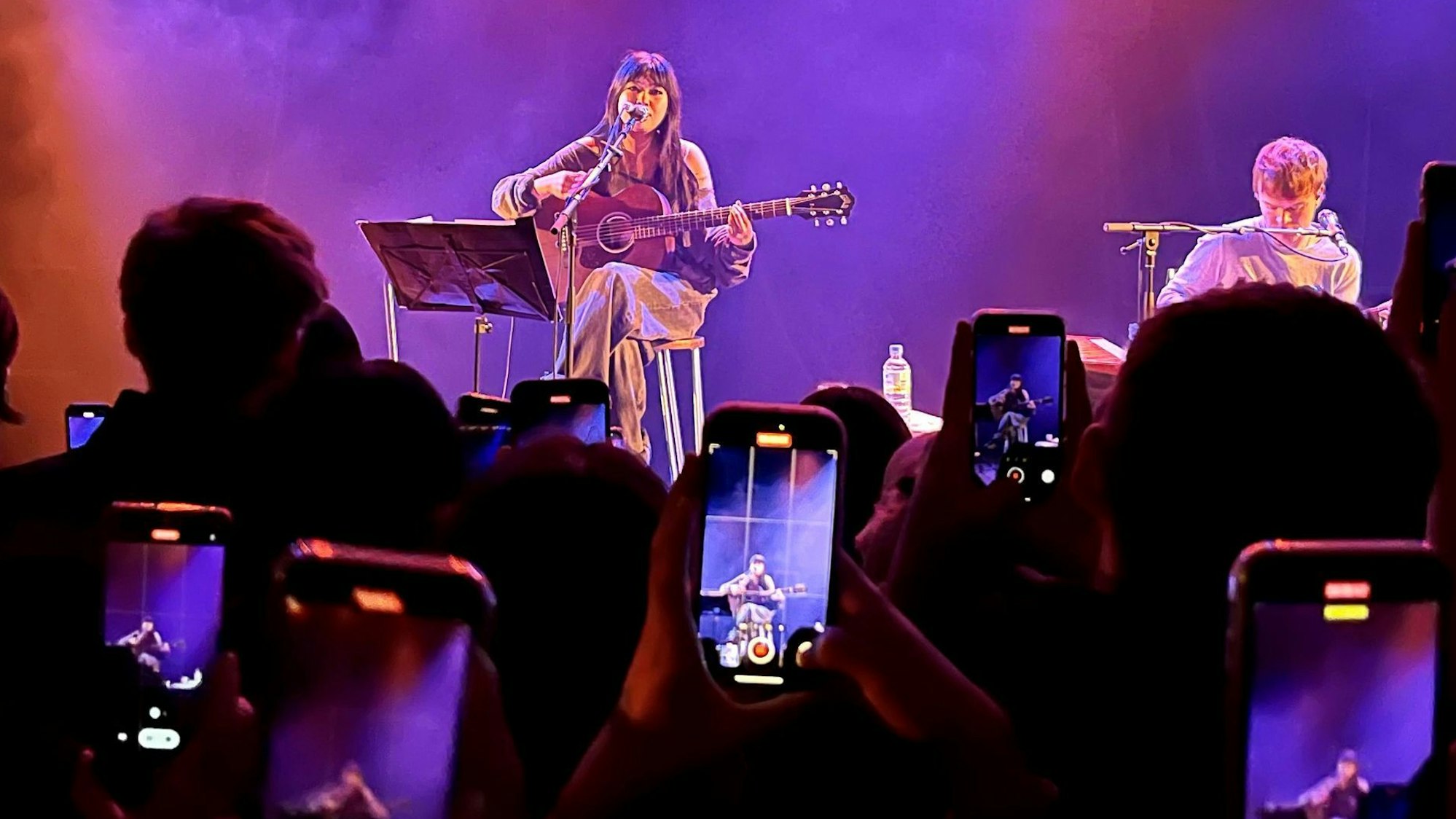 Die englische Sängerin Beabadoobee sitzt am 17. März 2023 mit einer Gitarre auf einem Barstuhl auf der Bühne des Kölner Bürgerhaus Stollwercks. Ihre Fans filmen sie mit ihren Handys ab.