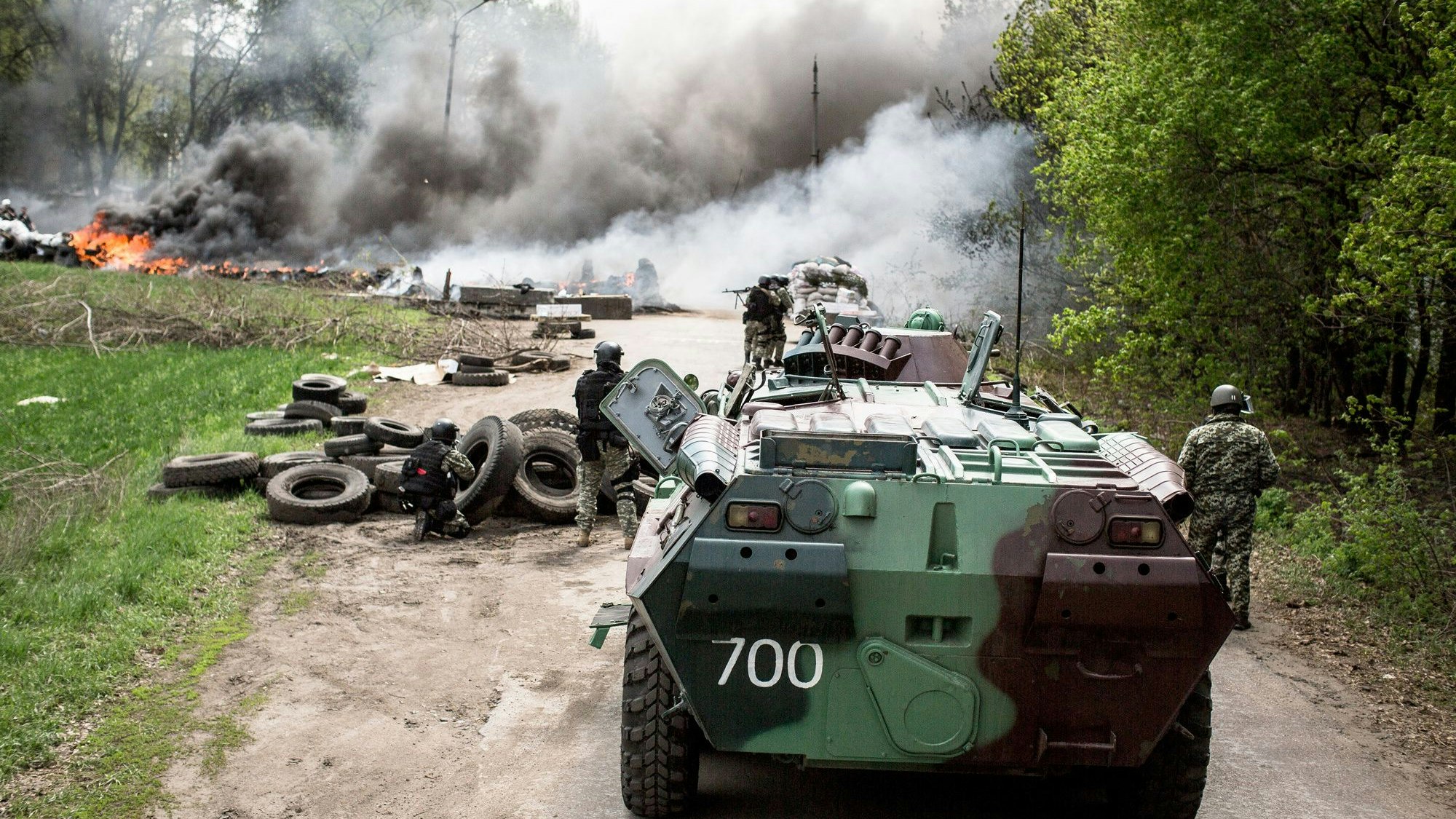 Ein Schützenpanzer steht auf einer Straße. Im Hintergrund sind Rauchwolken, Feuer und Gebäude zu sehen.