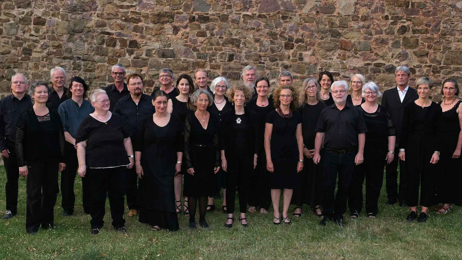 Etwa dreißig in schwarz gekleidete Personen stehen in drei Reihen aufgebaut vor einer Mauer.