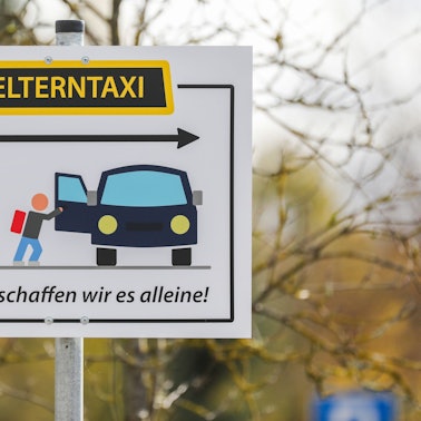 Ein Schild mit der Aufschrift „Elterntaxi Ab hier schaffen wir es alleine!“ steht am Parkplatz eines Schulgeländes.