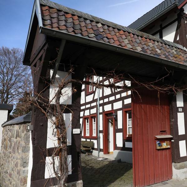 Über dem offenen Portal, das zu einem kleinen Hof führt, ranken Weinreben. Das Fachwerkhaus hat rote Fenstereinfassungen.