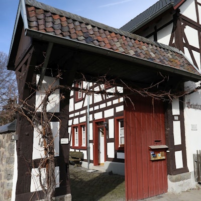 Über dem offenen Portal, das zu einem kleinen Hof führt, ranken Weinreben. Das Fachwerkhaus hat rote Fenstereinfassungen.