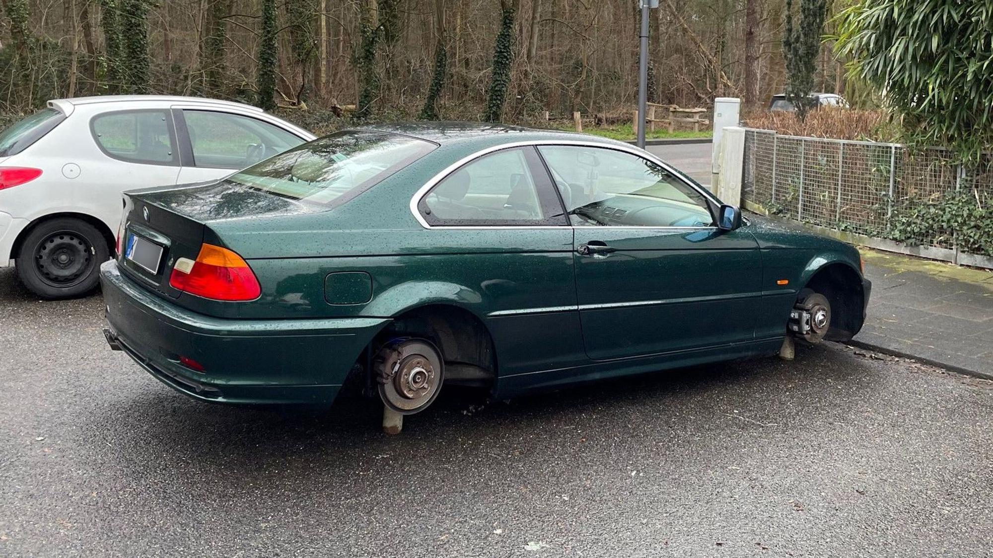 Ein schwarzer BMW steht mit demontierten Rädern auf Holzklötzen auf einem Parkplatz an der Straße „Am Klausenberg“ in Köln-Brück.