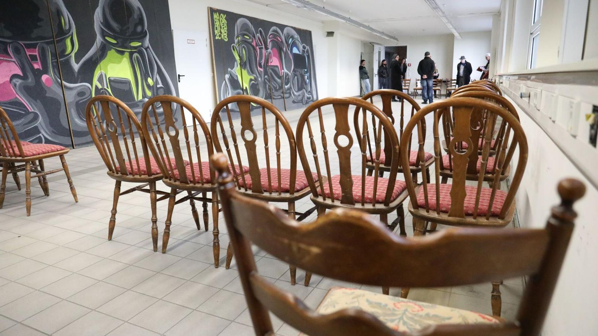 Im Vordergrund stehen in der ehemaligen Fabrikhalle braune Stühle, links an der Wand lehnen großformatige Streetart-Kunstwerke.