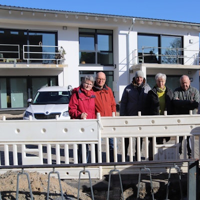 Mehrere Menschen stehen vor einem Mehrfamilienhaus. Vor ihnen befindet sich eine Baustelle.