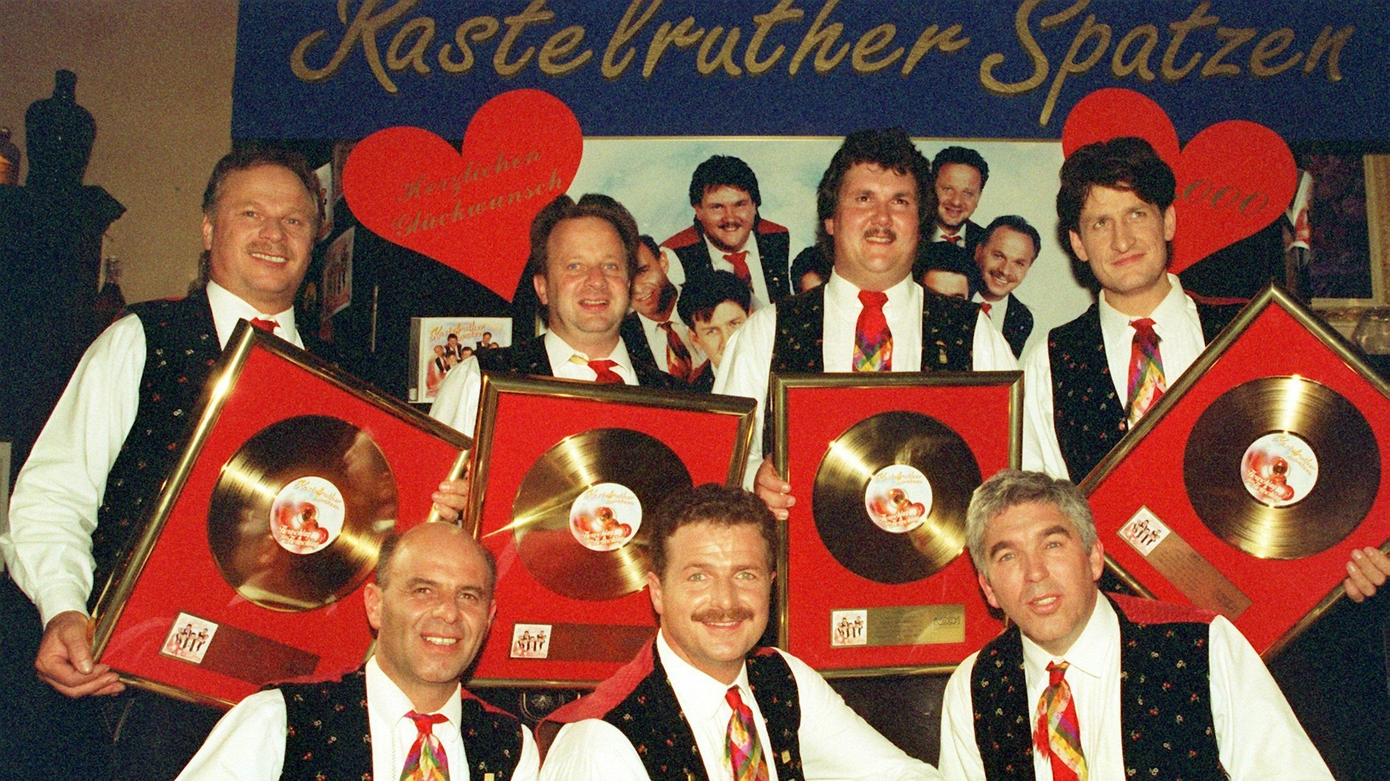 Die Kastelruther Spatzen mit Goldenen Schallplatten, Hinter ihnen ein Schild mit Herzen.