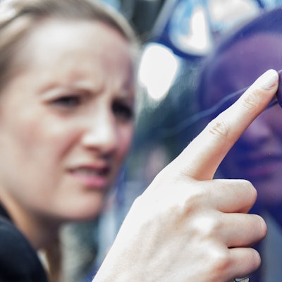 Eine Frau begutachtet an einen langen Kratzer im Lacke eines blauen Autos.