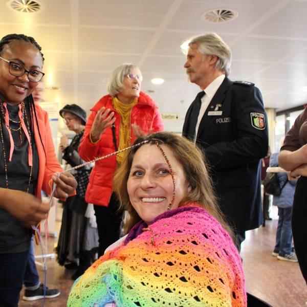 Epiphana Uwimana dreht einer in Regenbogenfarben gekleideten Besucherin bunte Fäden in die Haarsträhnen.