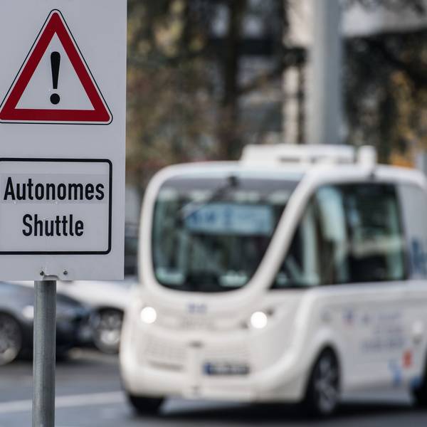Ein Schild "Autonomes Shuttle" weist auf dem Frankfurter Flughafen auf den Testbetrieb mit selbstfahrenden Shuttles hin, das im Hintergrund fährt.