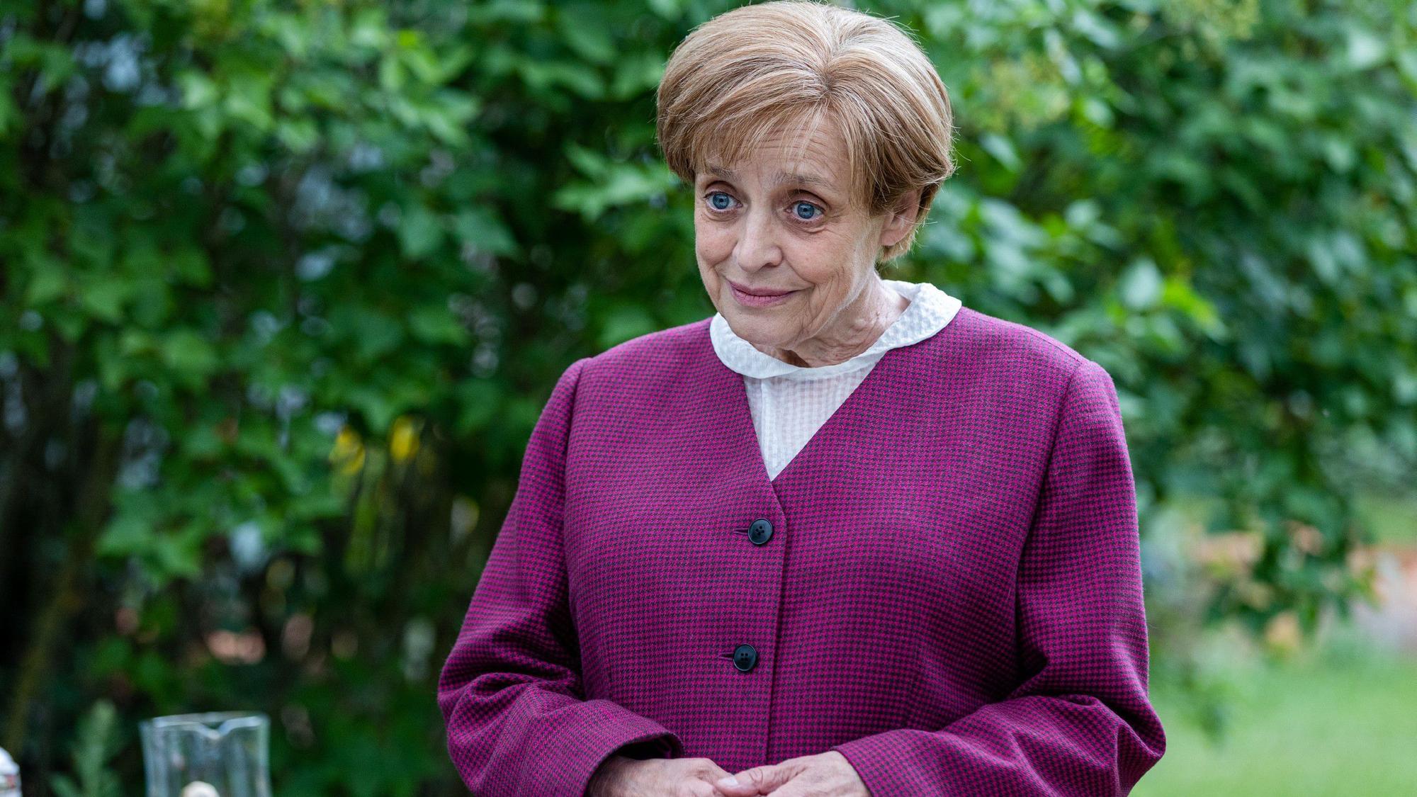 Miss Merkel (Katharina Thalbach) trägt einen violetten Blaser, sie hält die Hände in einer für die Kanzlerin typischen Geste vor sich.