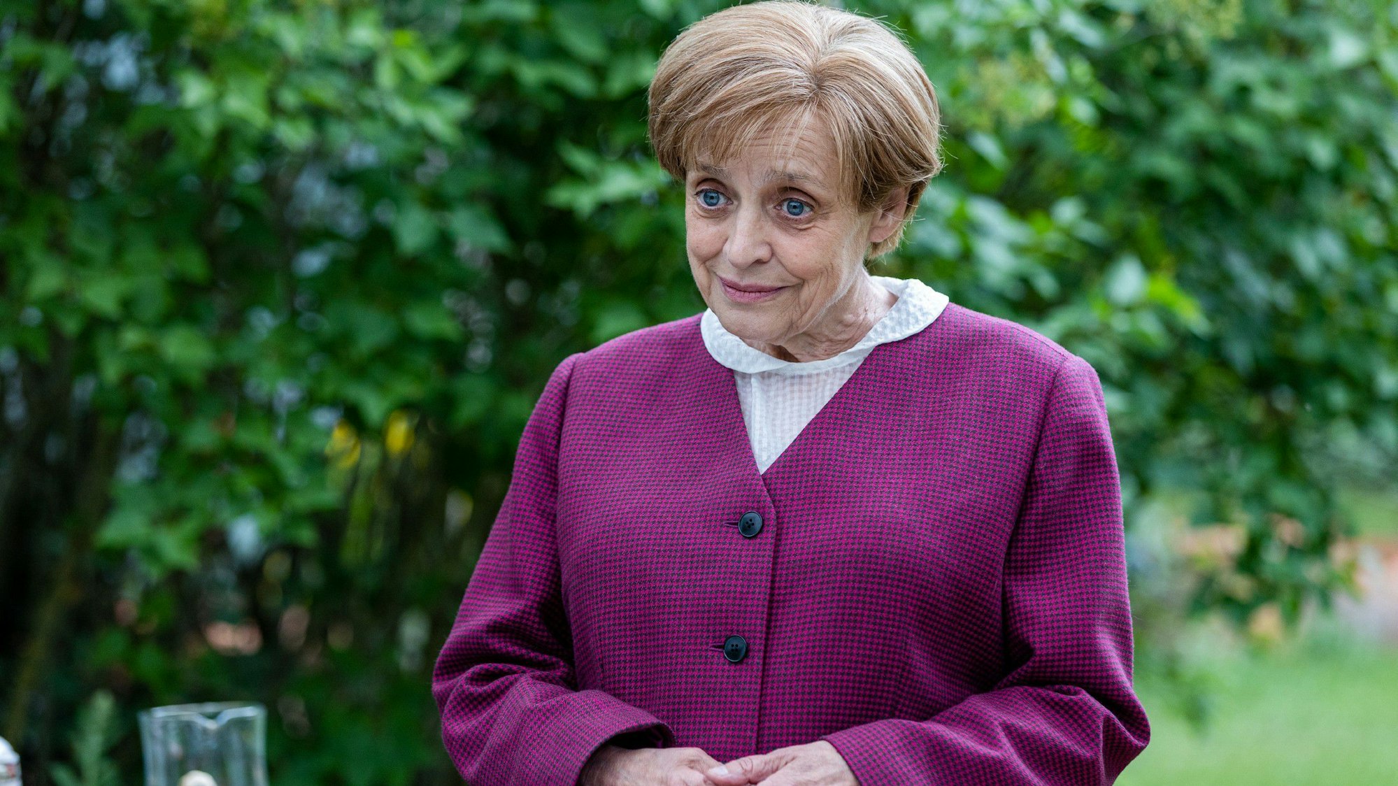 Miss Merkel (Katharina Thalbach) trägt einen violetten Blaser, sie hält die Hände in einer für die Kanzlerin typischen Geste vor sich.