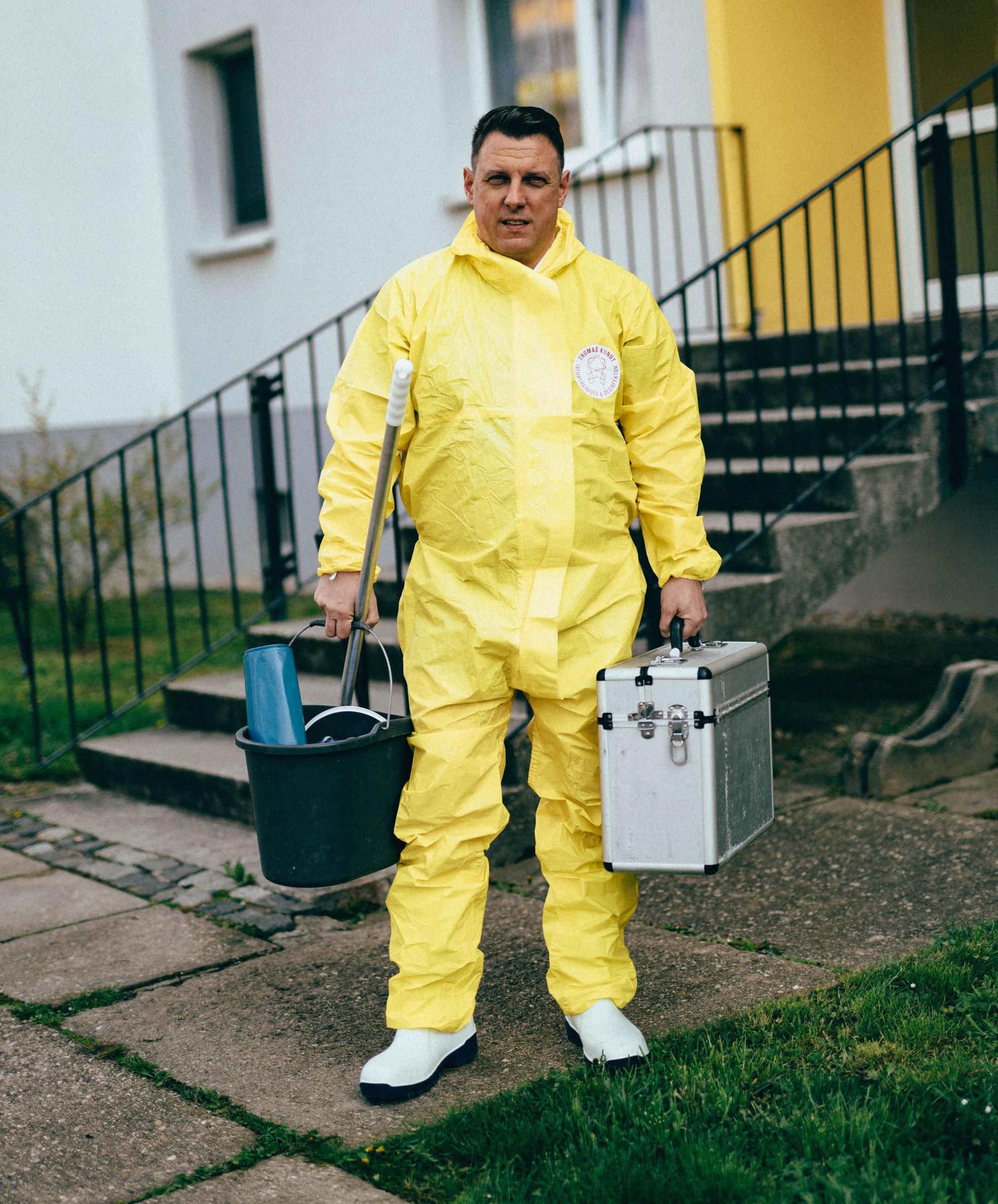 Thomas Kundt steht vor einem Haus. Er trägt einen gelben Schutzanzug und weiße Schutzschuhe. In den Händen trägt er Putzutensilien und einen Werkzeugkoffer.