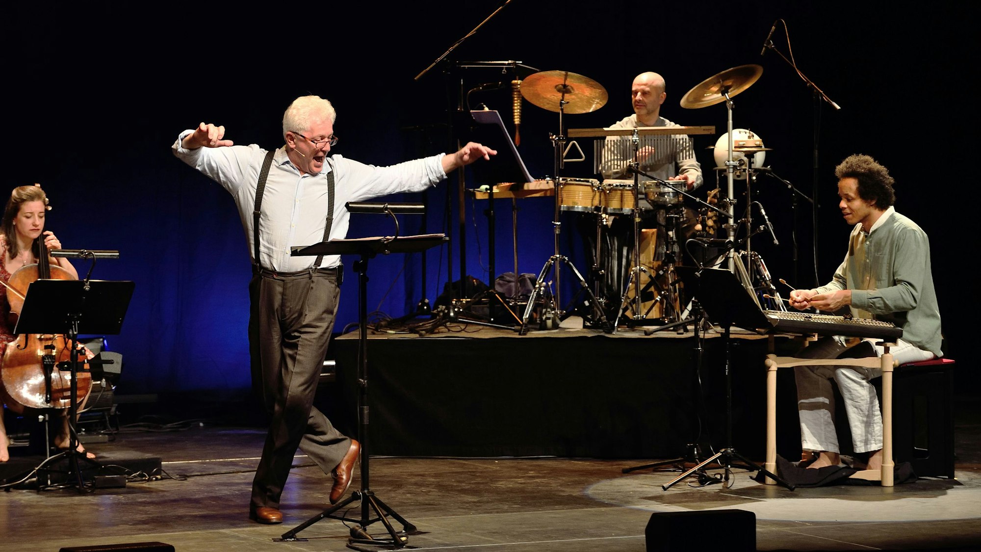 Miroslav Nemec steht zwischen Musikerinnen und Musikern auf der Bühne. Er tanzt mit ausgestreckten Armen.