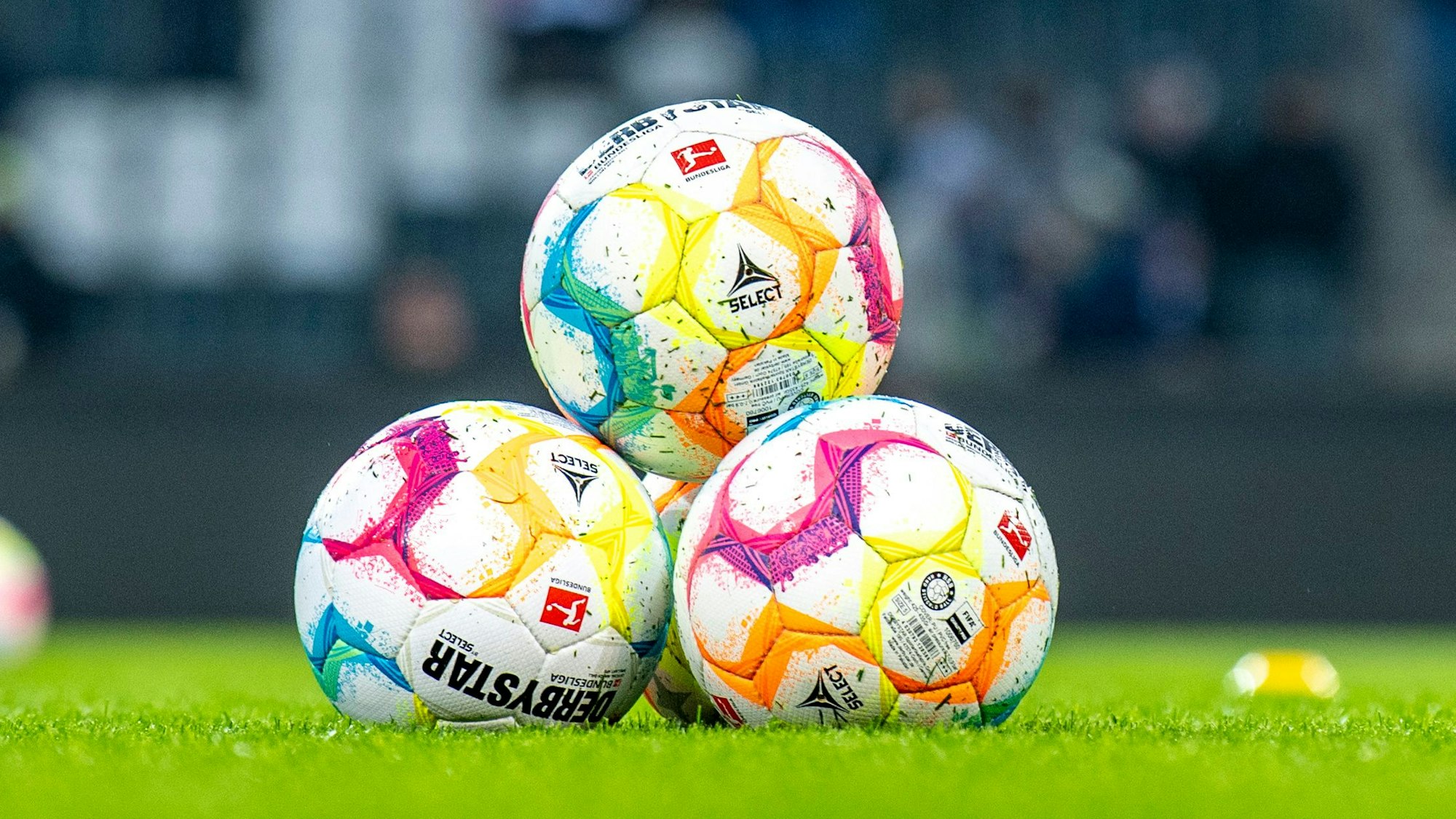 Vier Fußbälle liegen aufeinander gestapelt auf einem Rasen, im Hintergrund ist unscharf eine Tribüne zu erkennen.