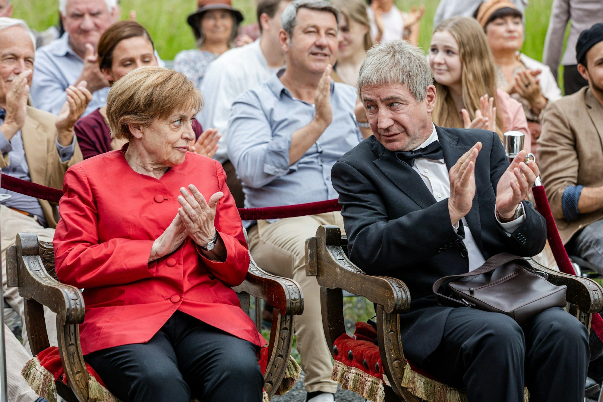 Das Ehepaar schaut sich an, beide klatschen. Die Kanzlerin trägt einen roten Blazer und eine schwarze Hose.