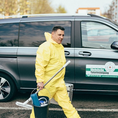 Zu sehen ist Thomas Kundt, der einen gelben Schutzanzug trägt und Putzutensilien mit sich hat. Im Hintergrund ist ein großes Auto auf dem „Sächsische Tatortreinigung“ steht.
